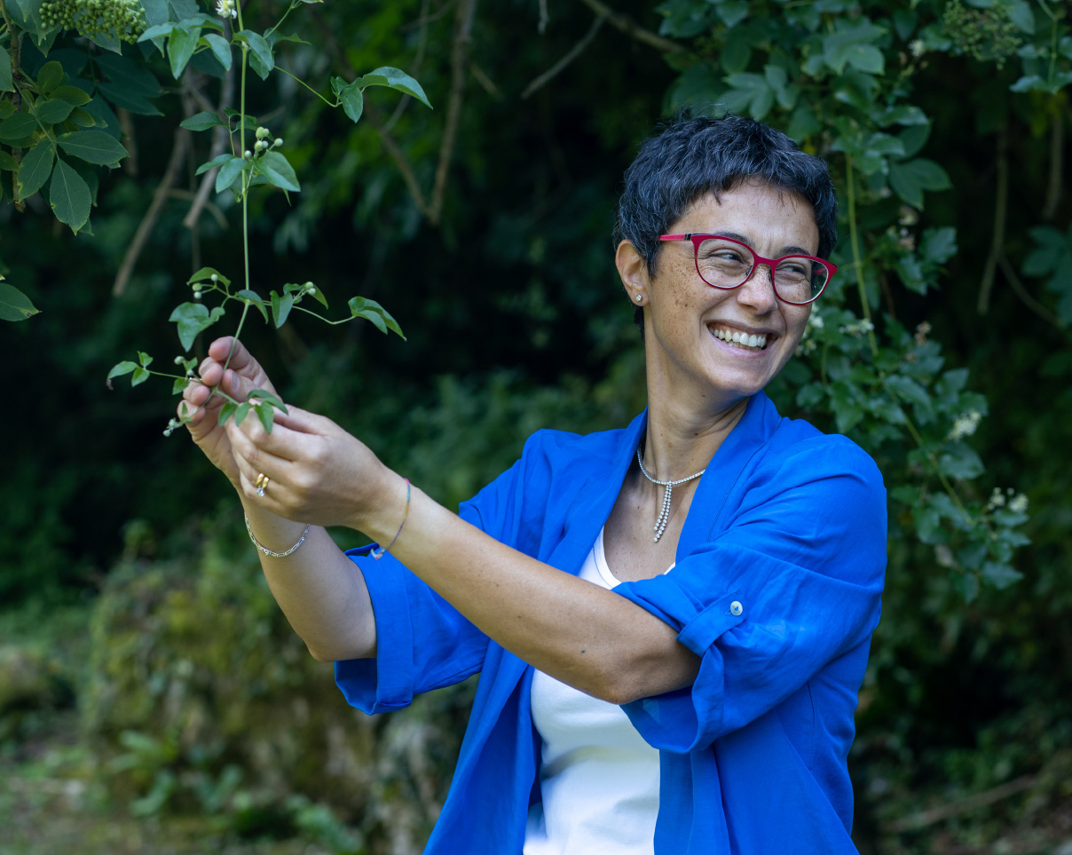 Foto di Nancy sorridente vestita di blu mentre tocca delle foglie di un albero immersa nella natura